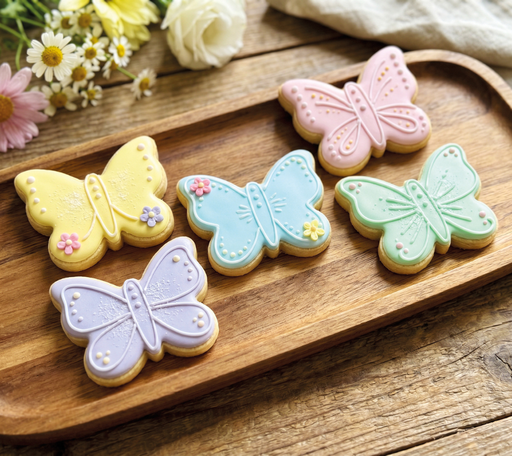 Auf einem Brettchen liegen 5 mit Pastellfarbenem Zuckerguss dekorierte Kekse in Schmetterlingsform